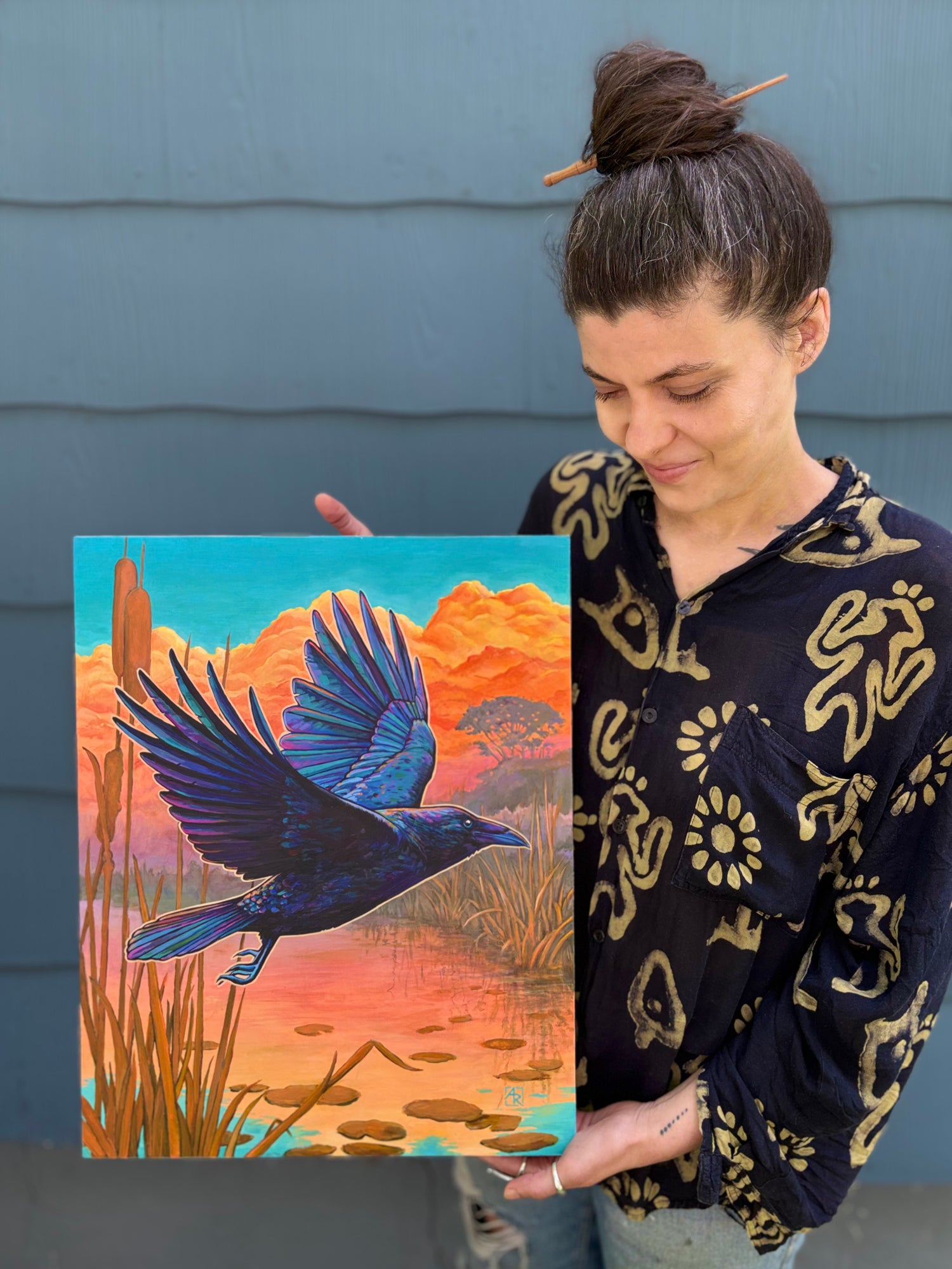 Woman holding a painting of a raven bird over water with a colorful landscape.