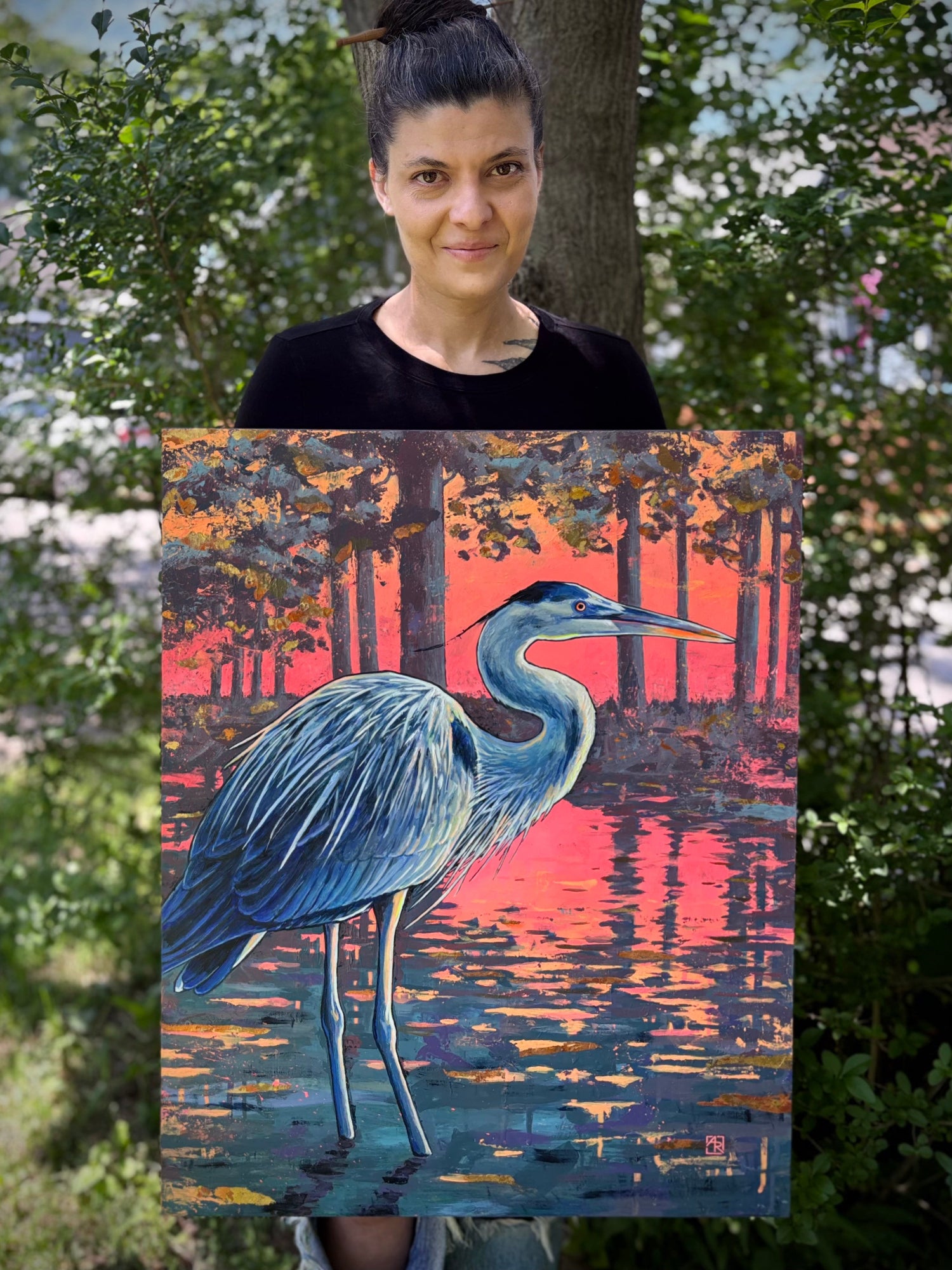 Person holding a painting of a heron at sunset with trees and water in the background.