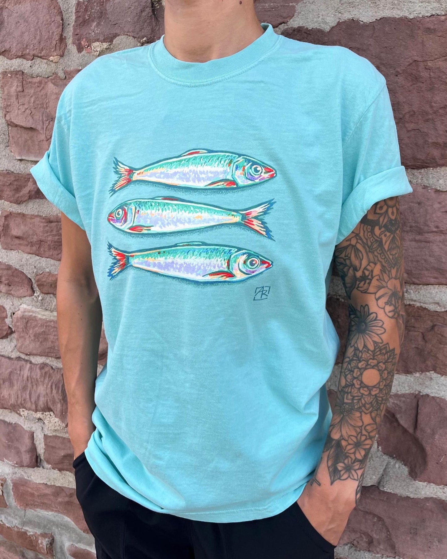 A person wearing a light blue t-shirt with a graphic of three sardine fish, standing against a brick wall.