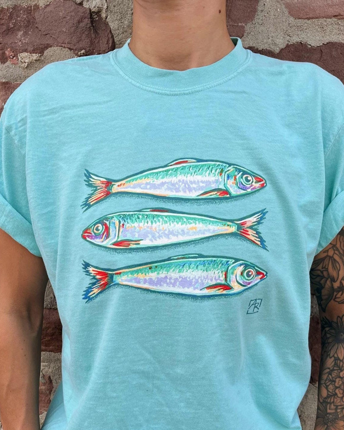 A person wearing a light aqua t-shirt with a graphic of three painted fish, standing in front of a rustic red brick wall.