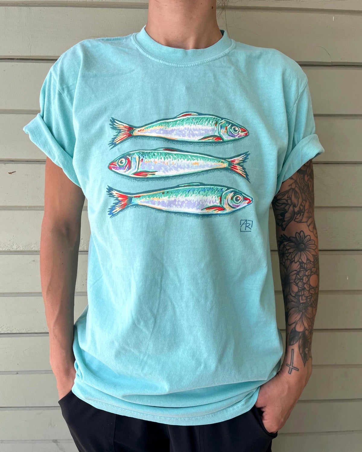 A person wearing a light aqua short-sleeve t-shirt with a graphic of three fish on the front, standing against a light wood siding background.